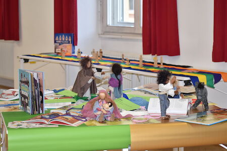 Tisch mit Puppen, Büchern und bunten Tüchern in einem hellen Raum.