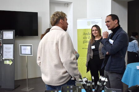 Drei Personen stehen und unterhalten sich bei einer Networking- oder Konferenzveranstaltung in einem Innenbereich.