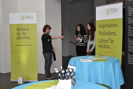 Drei Personen unterhalten sich neben Bannern und Tischen bei einer Bildungsveranstaltung oder Messe.