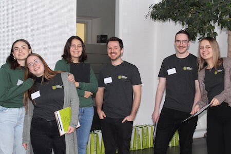 Sechs lächelnde Menschen stehen drinnen, manche halten Notizbücher und Papiere in den Händen.