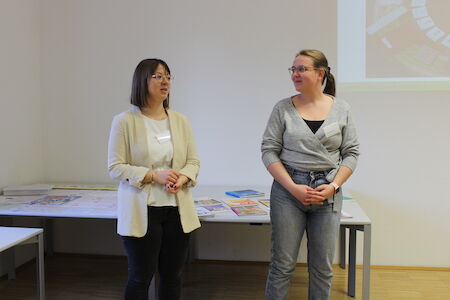 Zwei Frauen stehen und unterhalten sich in einem Klassenzimmer, hinter ihnen liegen Unterlagen auf den Tischen.