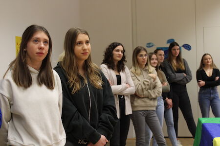 Eine Gruppe junger Frauen steht im Innenbereich und blickt aufmerksam auf etwas außerhalb des Bildes.