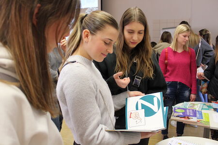 Drei junge Frauen schauen sich gemeinsam an einem Tisch mit weiteren Büchern ein aufgeschlagenes Buch an.