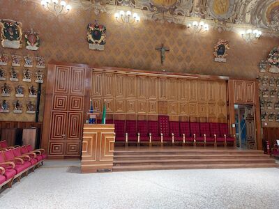 Reich verzierter Saal mit roten Stühlen, Holzvertäfelung, Wappen und einem Kreuz an der Wand.