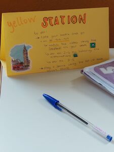 Eine gelbe Karte mit der Aufschrift „Gelbe Station“ liegt mit einem Stift und Papier auf einem Schreibtisch.