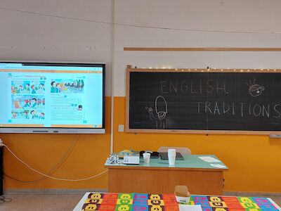 Klassenzimmer mit einem Smartboard, einer Tafel mit der Aufschrift „English Traditions“ und einem Schreibtisch.