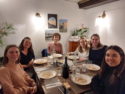 Sechs Frauen sitzen in einem gemütlichen Raum um einen Esstisch mit Tellern voller Essen und Getränken.
