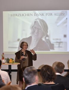 Eine Frau spricht bei einer Veranstaltung, während hinter ihr eine Dankesbotschaft projiziert wird.