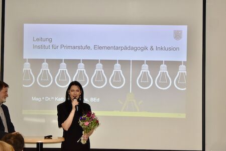 Eine Frau mit Blumen spricht am Podium, während hinter ihr eine Präsentation projiziert wird.