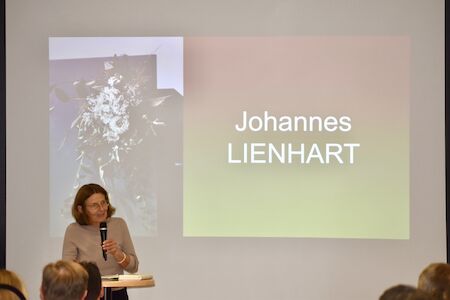 Eine Frau spricht an einem Podium vor einer Leinwand, auf der Johannes LIENHART zu sehen ist.