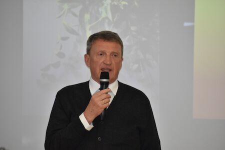 Mann im schwarzen Pullover spricht vor hellem Hintergrund in ein Mikrofon.