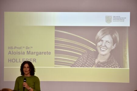Eine Frau spricht am Podium und zeigt auf einer Präsentationsfolie das Foto einer anderen Frau.
