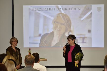Zwei Frauen stehen vor einem Bildschirm, eine hält Blumen und ein Mikrofon.