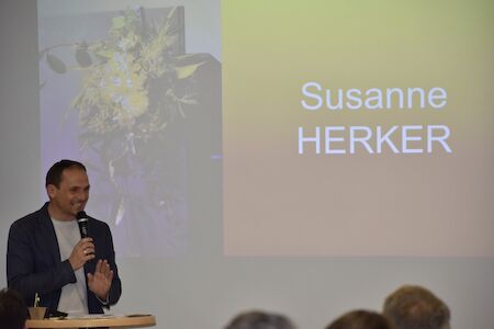 Mann spricht am Podium neben einem Bildschirm, auf dem Susanne HERKER zu sehen ist.