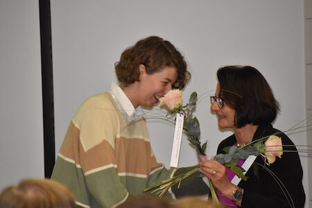 Zwei Frauen lächeln und tauschen Blumen bei einer Indoor-Veranstaltung aus.