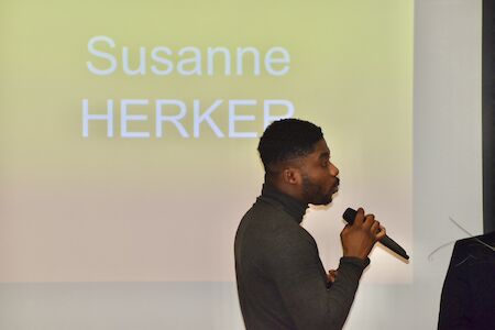 Mann spricht in ein Mikrofon vor einem Bildschirm, auf dem Susanne HERKER zu sehen ist.