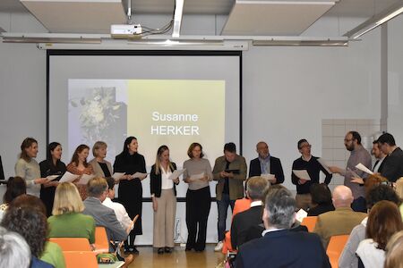 Eine Gruppe von Menschen steht und singt vor einem Publikum und einer Leinwand.