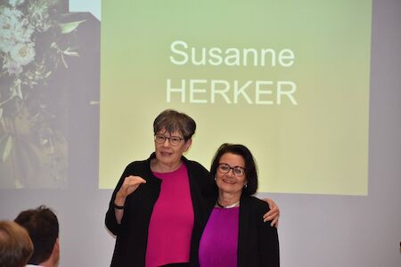 Zwei Frauen stehen zusammen vor einem Bildschirm mit der Aufschrift „Susanne HERKER“.