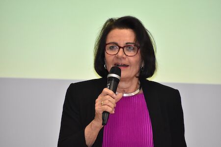 Frau mit Brille, die in ein Mikrofon spricht; sie trägt eine schwarze Jacke und ein lila Oberteil.