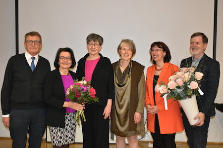 Sechs Erwachsene posieren im Innenbereich vor einem Bildschirm, zwei halten Blumensträuße in der Hand.