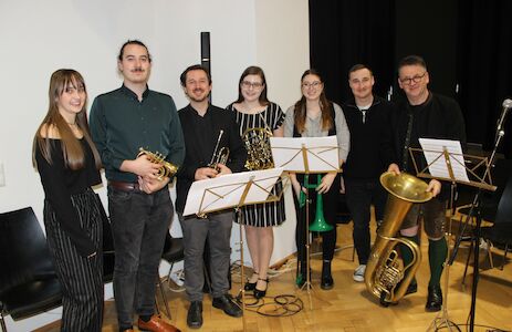 Sieben Musiker mit Blechblasinstrumenten stehen lächelnd zusammen in einem Musikzimmer.
