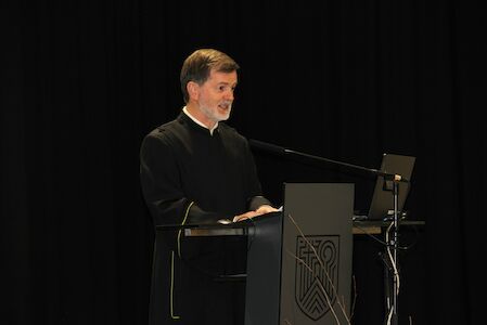 Ein Mann in einer schwarzen Robe spricht vor einem dunklen Hintergrund an einem Podium mit einem Laptop.