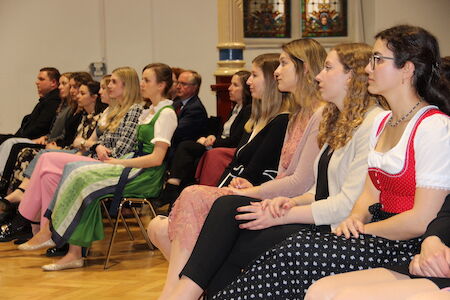 Menschen sitzen drinnen, einige Frauen tragen traditionelle Kleidung und schauen sich eine Veranstaltung an.