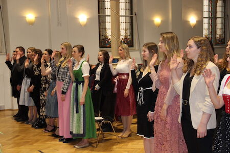 Eine Gruppe von Frauen steht mit erhobener rechter Hand bei einer formellen Zeremonie im Innenbereich.