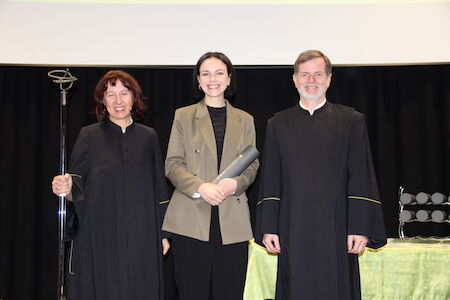 Drei Personen stehen zusammen, zwei in schwarzen Roben, eine in einem hellbraunen Blazer, alle lächeln.