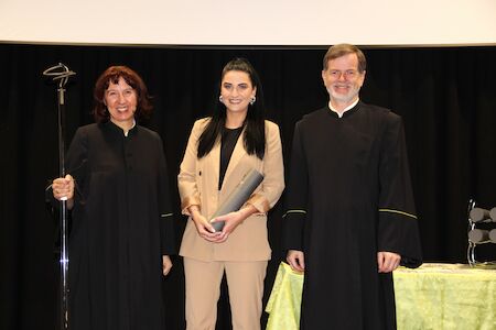 Drei Personen posieren bei einer Zeremonie; einer hält eine Schriftrolle, zwei tragen schwarze akademische Roben.