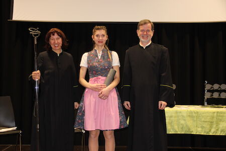 Auf der Bühne posieren drei Personen, zwei in schwarzen Gewändern und eine im rosa Dirndl.