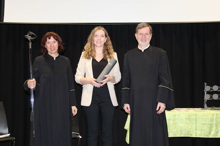 Drei Personen posieren; zwei in schwarzen Roben, einer im Blazer und mit einer Schriftrolle in der Hand.