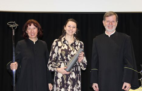 Drei lächelnde Menschen in schwarzen Roben stehen zusammen; einer hält eine Schriftrolle.