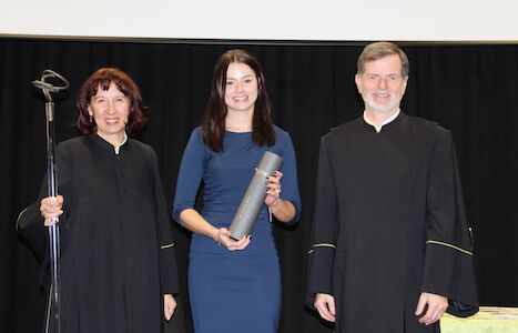 Drei Personen in schwarzer Abendkleidung; die Frau in der Mitte hält eine gerollte Urkunde.