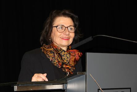 Frau mit Brille und gemustertem Schal spricht auf einem Podium vor dunklem Hintergrund.