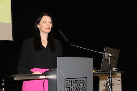 Eine Frau mit langen dunklen Haaren spricht an einem Podium, an dessen Seite ein Laptop steht.