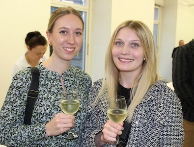 Zwei Frauen lächeln und halten Gläser mit Weißwein bei einer Veranstaltung in geschlossenen Räumen.