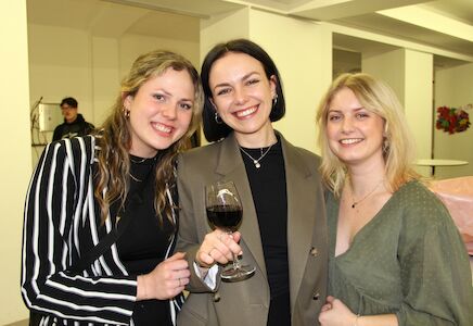 Drei lächelnde Frauen, eine davon mit einem Glas Rotwein in der Hand, drinnen bei einer Veranstaltung.
