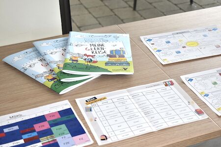 Auf einem Holztisch liegen Hefte mit dem Titel „Meine Lernreise“ und Arbeitsblätter aus.