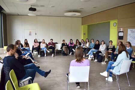 Menschen sitzen in einem großen Kreis und diskutieren in einem hellen Klassenzimmer.