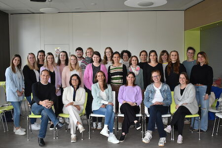 Eine Gruppe von 24 Personen posiert gemeinsam sitzend und stehend in einem modernen Raum.