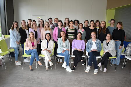 Eine Gruppe von 25 Personen posiert gemeinsam in einem Innenbereich, sitzend und stehend in Reihen.
