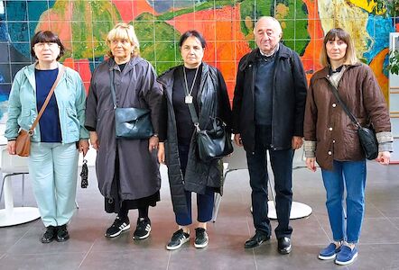 Fünf Erwachsene stehen im Innenbereich vor einer bunten Mosaikwand und blicken in die Kamera.