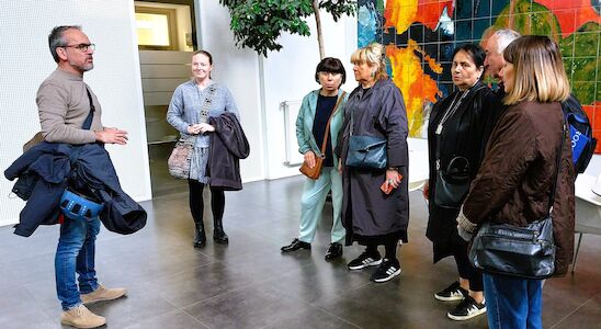 Ein Mann spricht in einem Innenbereich neben einer bunten Wandkarte zu einer Gruppe von sechs aufmerksamen Personen.