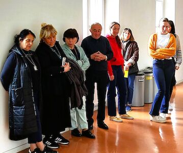 Sieben Personen stehen im Innenbereich an einer Wand, manche blicken in die Kamera, manche nicht.