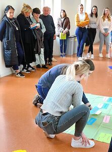 Zwei Frauen knien neben einer bunten Tafel, während andere drinnen stehen und zuschauen.