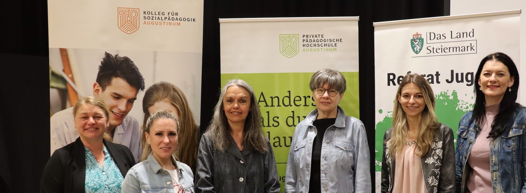 Sechs Frauen stehen und lächeln vor Bildungs- und Regierungsbannern.