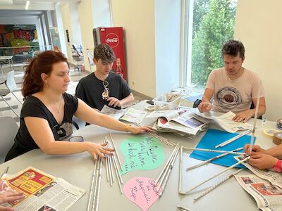 Vier Personen sitzen an einem Tisch und basteln mit Zeitungen und buntem Papier.