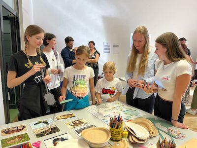 Kinder und Erwachsene versammeln sich um einen Tisch mit Bastel- und Künstlerbedarf.