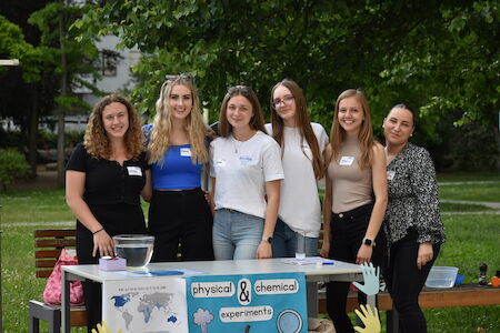 Sechs junge Frauen stehen hinter einem Tisch mit der Aufschrift „Physikalische und chemische Experimente“.